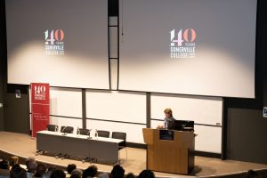 Vice Chancellor Louise Richardson stands on stage speaking at the lectern, with the somerville 140 logo projected above