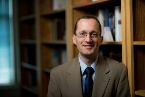 Albert Pionke, wearing a suit and glasses, sits in front of a bookcase.