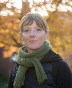 Sophie Walwin is wearing an olive green scarf and a black gilet and stands in front of a tree with orange autumnal leaves in Somerville's quadrangle