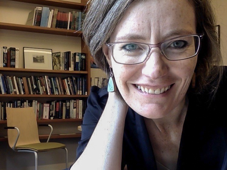 Professor Patricia Owens, a woman with glasses, brown hair, and blue earrings, smiles to the camera. Behind her is a bookcase and a chair in her office. 