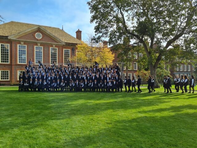 Undergraduate students file in to a temporary stand set up on the quad lawn for their yeargroup matriculation photo. They are dressed smartly in their sub fusc, with academic gowns and white and black bow ties or black ribbons. Around them, the quadrangle looks beautiful on a sunny autumn morning.