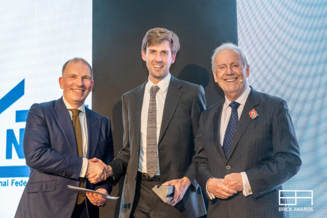 Alistair browning, a tall young architect in a dark suit, is holding his Brick Award, a small concrete model of a brick house, and using his other hand to shake hands with a member of the panel, a man in a dark suit, to his right. On his left is Gyles Brandreth, a man in a dark suit in his 70s. They are all beaming.