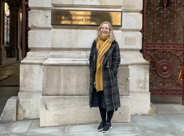 Dr Albury stands outside the Foreign, Commmonwealth and Development Office in Whitehall underneath their brass sign, looking very excited to be there. She has blonde hair, metal-rimmed glasses, a yellow scarf and a warm woollen tartan winter coat.