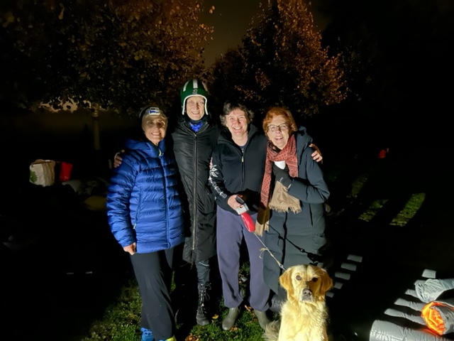 Maggie Snowling, Sarah Springman, Irene Tracey, and Jan Royall are standing next to eachother in their warm coats and hats. Even though it's dark and cold, they are smiling, glad to be together and doing something to raise money and awareness. Irene has her dog with her, a golden retriever who is sat very patiently in front of them doing their best serious pose.