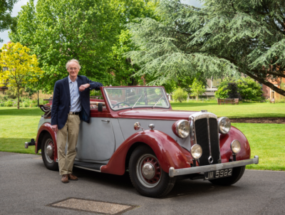 A man stands proudly next to a beautiful vintage car in a sunny quad.