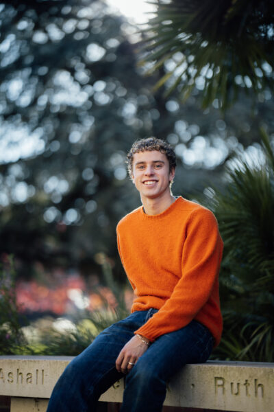 A young white man in an orange jumper sits on a bench and smiles at the camera