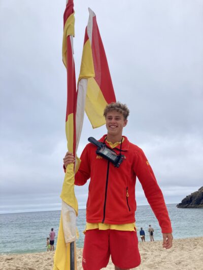 A smiling young man in red and yellow lifeguard clothes holds up lifeguarding flags on a beach