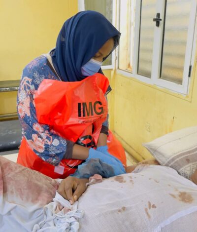 A young woman wearing a hijab and protective orange clothing stands over the bedside of an injured man, attending to his wounds