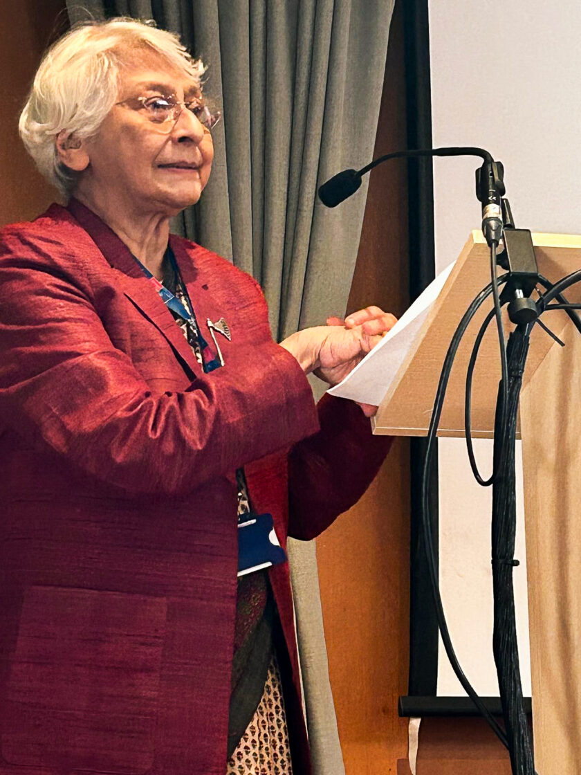 A distinguished woman dressed in a smart red jacket stands at a microphone