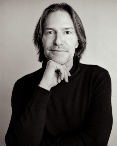 A man looks straight into the camera, with a calm, serious expression and a slight smile. The photograph is in black and white.