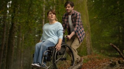 A man pushes a woman in a wheelchair through a forest, both look frightened.